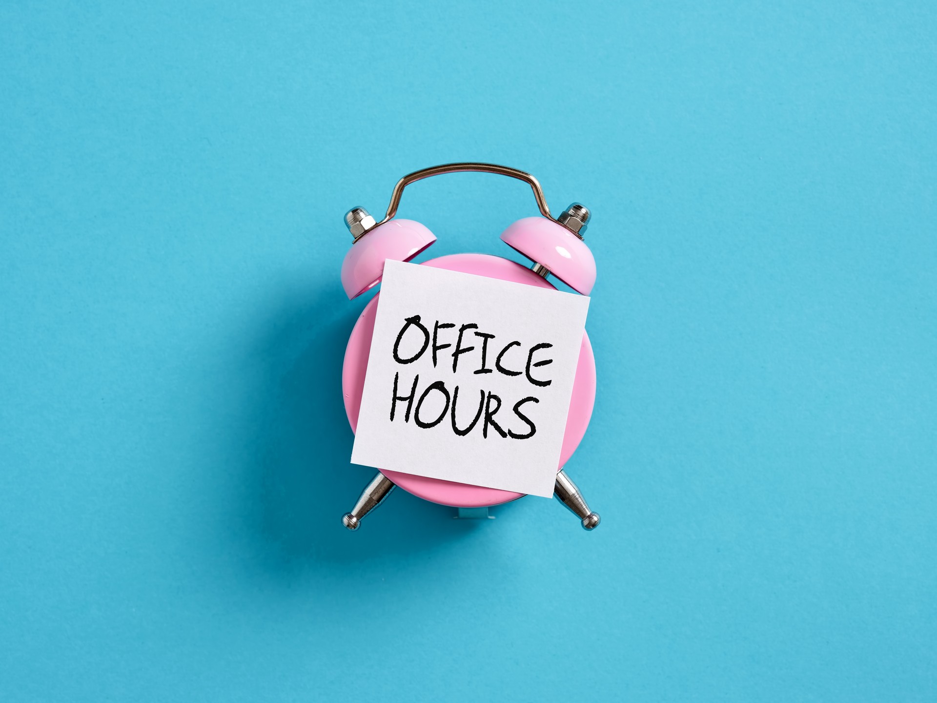 Office hour concept in business or education. Pink alarm clock with a note paper on blue background with the word office hours.